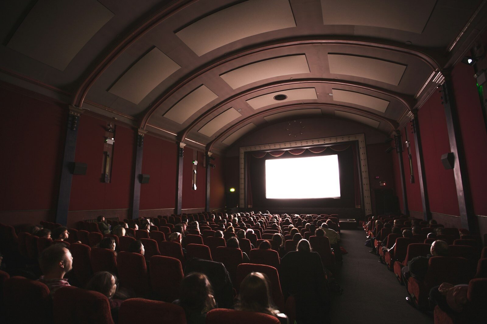 An audience in a dark cinema with red velvet seats facing a bright screen