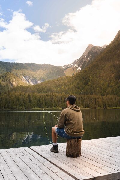 man in brown t-shirt and blue shorts standing on dock looking at lake during daytime