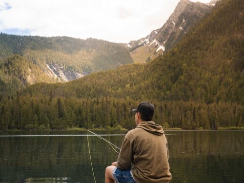 man in brown t-shirt and blue shorts standing on dock looking at lake during daytime