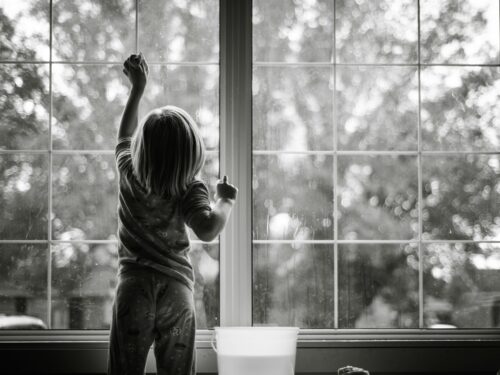 grayscale photo of girl in stripe shirt and pants standing by the window