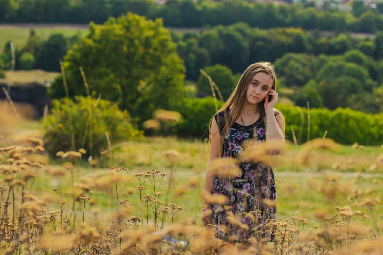 A young woman in a floral dress stands thoughtfully in a grassy field on a sunny day.