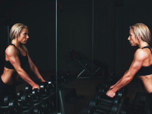 woman wearing black top top holding black dumbbells standing in front of mirror