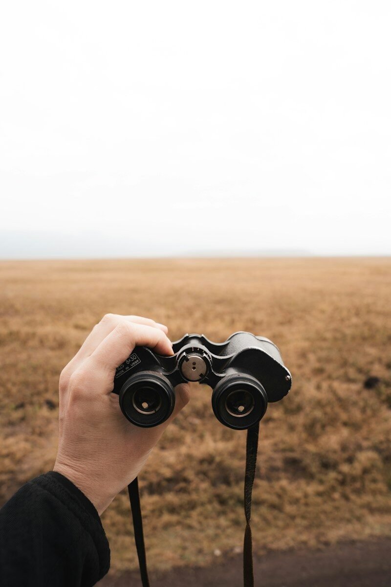 A hand holds binoculars, overlooking a field