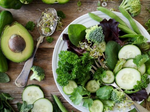 sliced broccoli and cucumber on plate with gray stainless steel fork near green bell pepper, snowpea, and avocado fruit