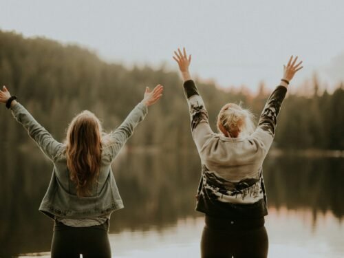 two women hands up standing beside body of water